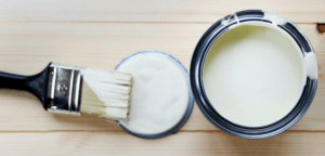 An open paint can sits on a wooden table with a paint-covered brush on the upturned lid.
